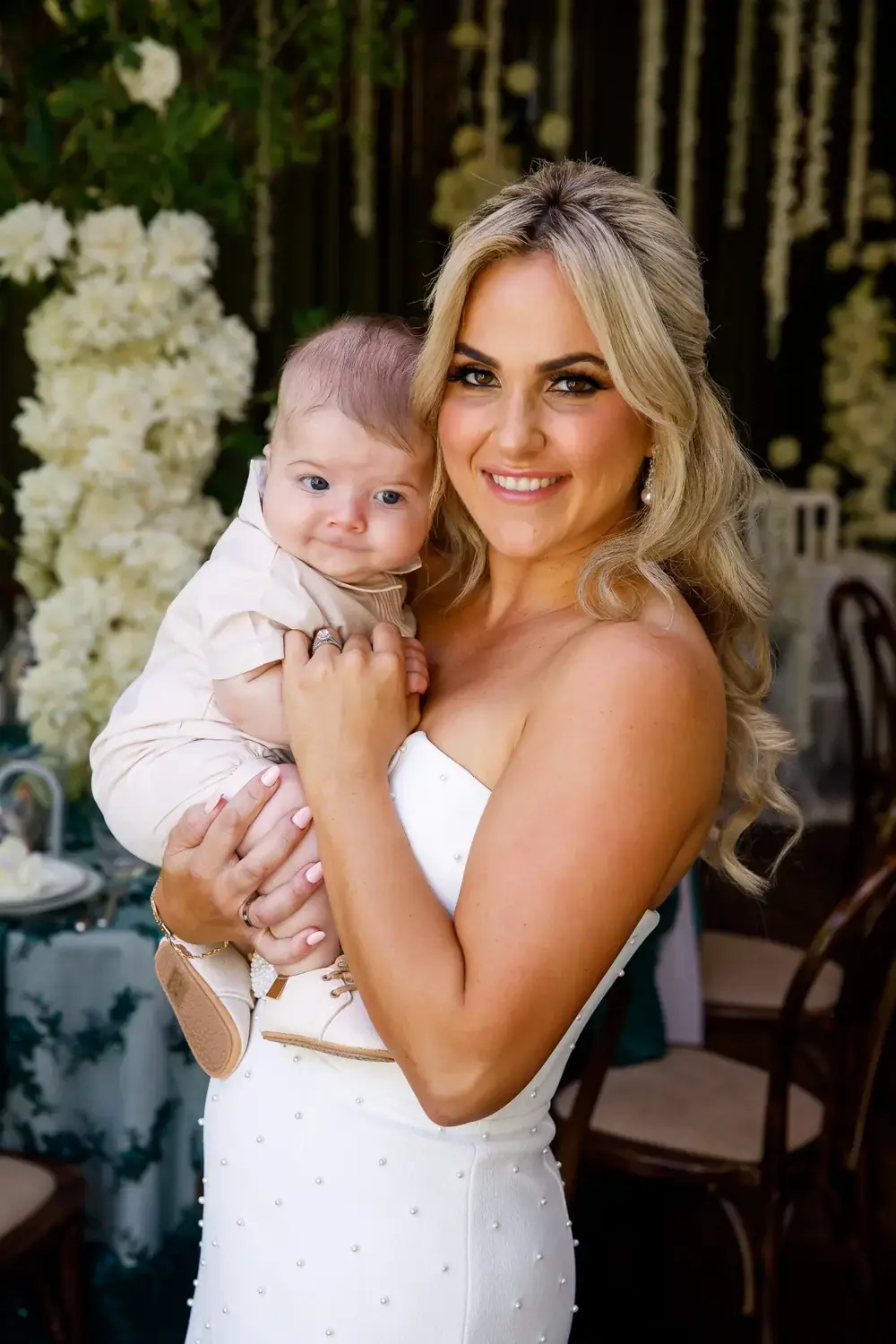 Elegant christening floral styling with mother holding baby surrounded by white roses and greenery